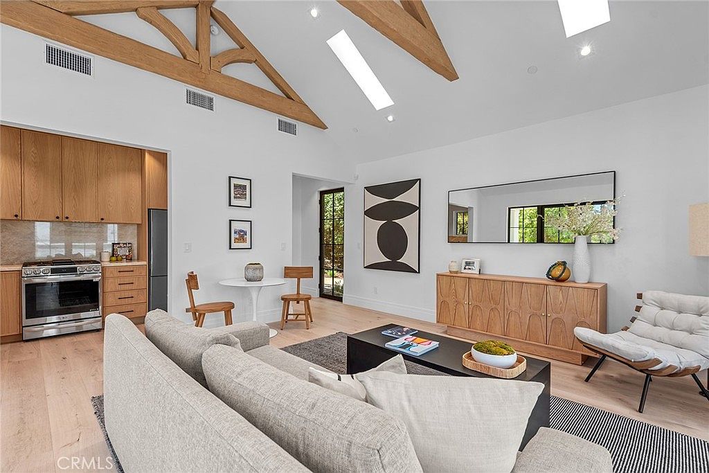 This bright and airy open-concept living space features vaulted ceilings with exposed wooden beams and light-toned hardwood flooring. The room is anchored by a comfortable grey sofa, a sleek black coffee table, and a modern accent chair, all complemented by minimalist decor and a large wall mirror. The seamless transition into the adjacent kitchen area creates a spacious, inviting atmosphere perfect for modern living.