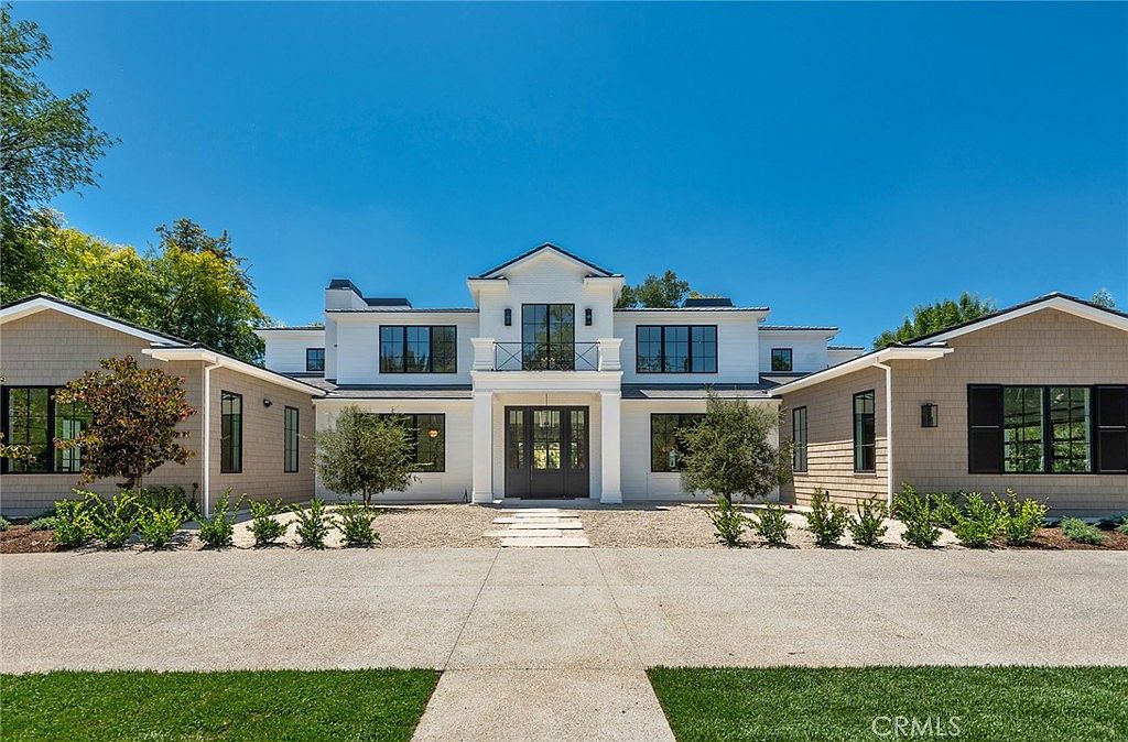 This stunning modern farmhouse features a symmetrical facade with a crisp white exterior contrasted by warm, light-brown shingle siding on the wings. The grand entryway is framed by a central gable and a second-story balcony, while large, dark-framed windows provide a sophisticated, balanced aesthetic. A clean, gravel-lined path leads to the front door, set against a backdrop of lush green trees and a clear blue sky, creating an inviting and luxurious curb appeal.