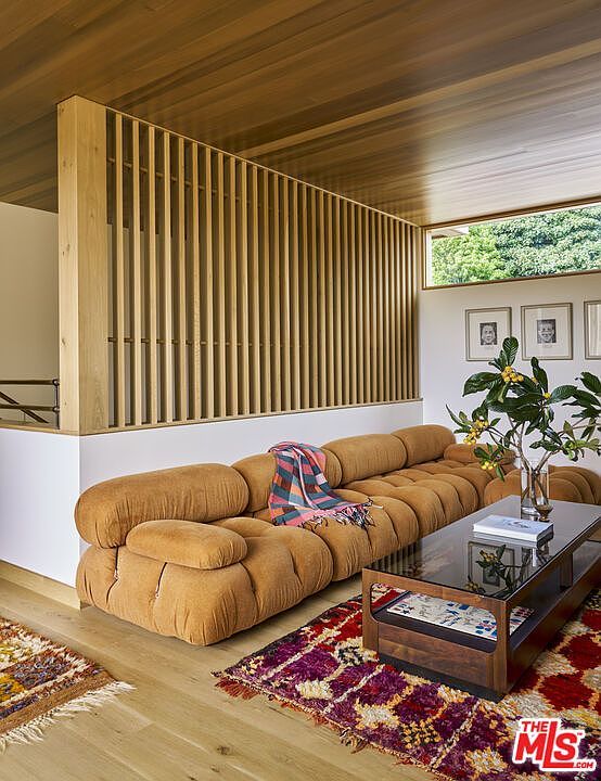This living room features a distinctive, textured modular sofa in a warm, earthy tone, positioned against a striking vertical wood slat room divider. The space is accented by a vibrant, patterned area rug and a low-profile coffee table with a glass top, while a large potted plant adds a touch of greenery. The room is bathed in natural light from a horizontal window, creating a cozy and sophisticated atmosphere with a mid-century modern aesthetic.