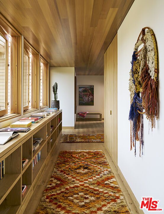 This warm and inviting hallway features a long, built-in light wood bookshelf lining one wall, topped with a collection of books and a potted cactus. The space is accented by a textured, colorful wall hanging and a plush, patterned runner rug that leads the eye toward a secondary seating area in the distance. With its natural wood-paneled ceiling and soft, ambient lighting, the hallway exudes a cozy, bohemian-inspired aesthetic.