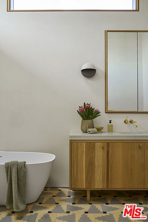 This modern bathroom features a freestanding white soaking tub adorned with a soft green towel, positioned next to a light wood vanity with a stone countertop. A gold-toned faucet and a matching framed mirror add a touch of elegance, while a unique wall-mounted light fixture provides soft illumination. The space is grounded by a striking patterned tile floor with geometric shapes, creating a clean and sophisticated aesthetic.