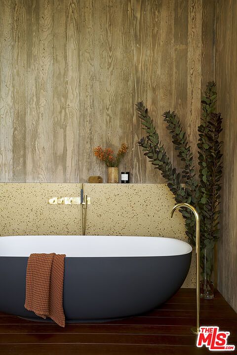 This sophisticated bathroom features a deep, matte charcoal soaking tub set against a backdrop of textured wood-look wall paneling and a speckled stone ledge. A sleek, gold-toned floor-mounted faucet complements the minimalist aesthetic, while a tall vase of greenery adds a natural, organic touch. The warm lighting and rich materials create a serene, spa-like atmosphere.