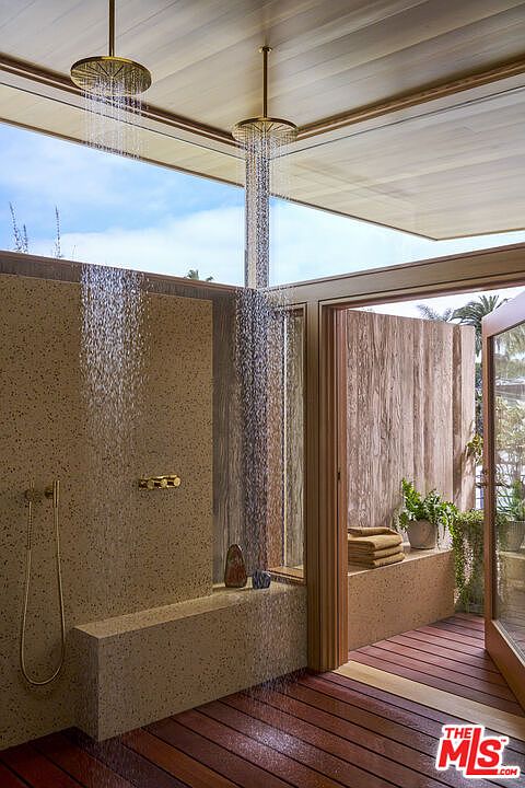This luxurious indoor-outdoor shower features dual rainfall showerheads suspended from a light wood-paneled ceiling. The space is defined by textured, speckled stone walls and a matching built-in bench, complemented by warm-toned wood decking. A glass door opens to an adjacent outdoor area, creating a seamless transition between the shower and the exterior environment.