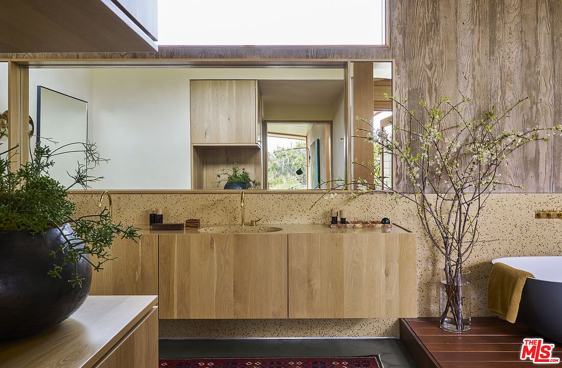 This sophisticated primary bathroom features a floating light wood vanity with a speckled stone countertop and integrated sink. A large, wide mirror reflects the room, while a textured wood-paneled wall adds warmth and organic character to the space. To the right, a dark soaking tub sits on a raised wooden platform next to a vase of flowering branches, creating a serene, spa-like atmosphere.