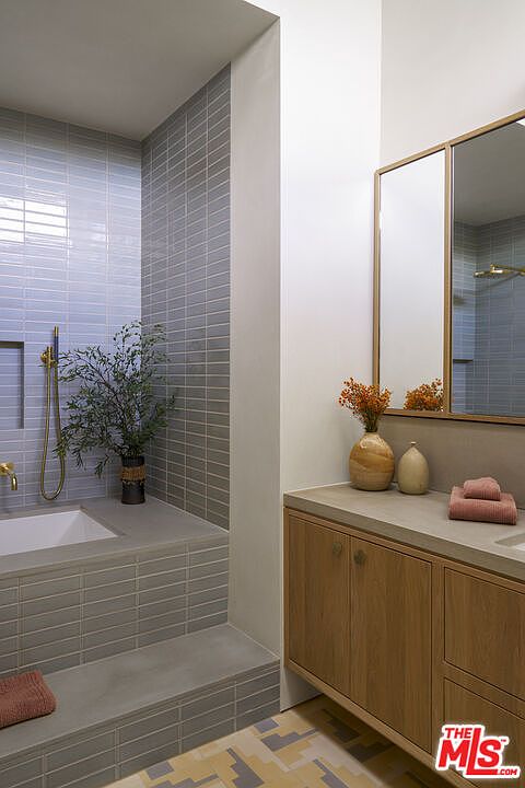 This modern bathroom features a built-in soaking tub surrounded by light-colored, rectangular wall tiles and a concrete-style ledge. To the right, a light wood vanity with a stone countertop is topped by a mirror and decorative vases, while the floor showcases a geometric, multi-colored tile pattern. The space is bright and minimalist, emphasizing clean lines and a neutral color palette.
