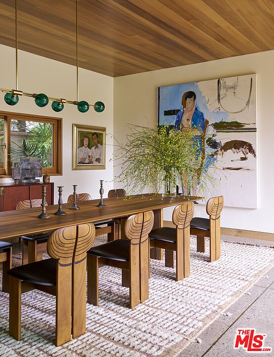 This sophisticated dining room features a long wooden table surrounded by unique, sculptural chairs with curved, organic backrests. A modern linear pendant light with green glass globes hangs above the table, while a large, vibrant abstract painting serves as a focal point on the wall. The space is grounded by a textured area rug and accented by a wood-paneled ceiling, creating a warm and artistic atmosphere.