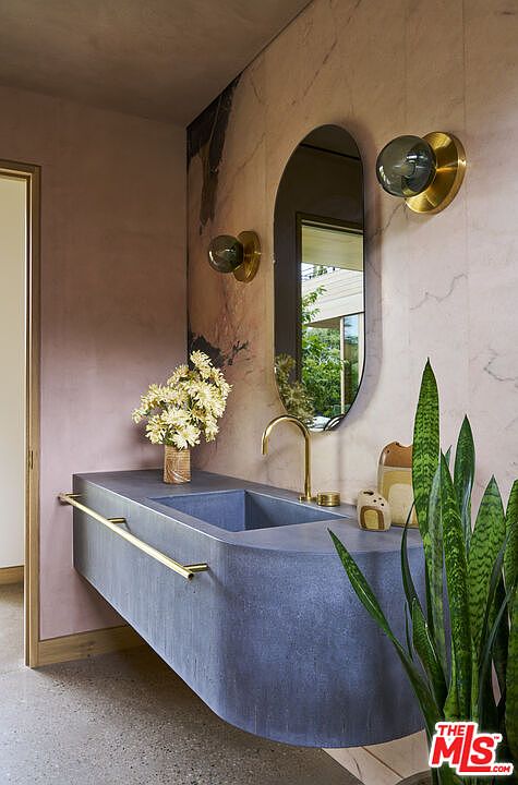 This sophisticated powder room features a striking, curved, blue-toned vanity with a gold-toned faucet and a matching towel bar. The wall behind the vanity is adorned with a large, oval mirror flanked by two globe-style wall sconces, set against a textured, blush-colored wall. A tall snake plant in the foreground and a vase of flowers on the vanity add organic elements to this modern, high-design space.
