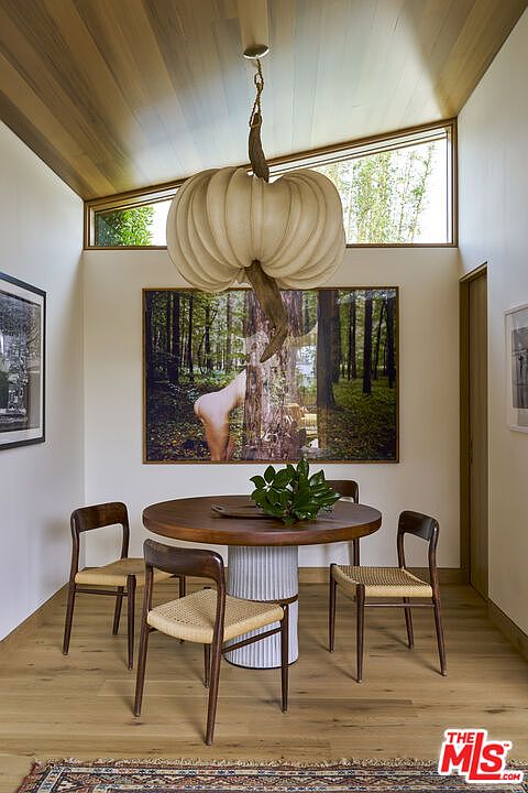 This elegant dining area features a round wooden table centered beneath a unique, sculptural pendant light fixture. The room is framed by a vaulted wood-paneled ceiling and a large, wide window that allows natural light to fill the space. A large piece of wall art serves as a focal point, complemented by four mid-century modern style chairs with woven seats, all set upon light-toned wood flooring.