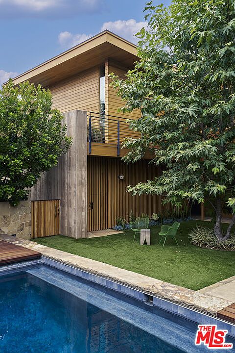 This contemporary residence features a striking blend of vertical wood siding and a prominent concrete structural element, creating a modern architectural aesthetic. The backyard includes a serene swimming pool in the foreground, a manicured lawn area with two green chairs, and a mature tree providing natural shade. The composition captures a private, tranquil outdoor living space that seamlessly integrates natural textures with clean, geometric lines.