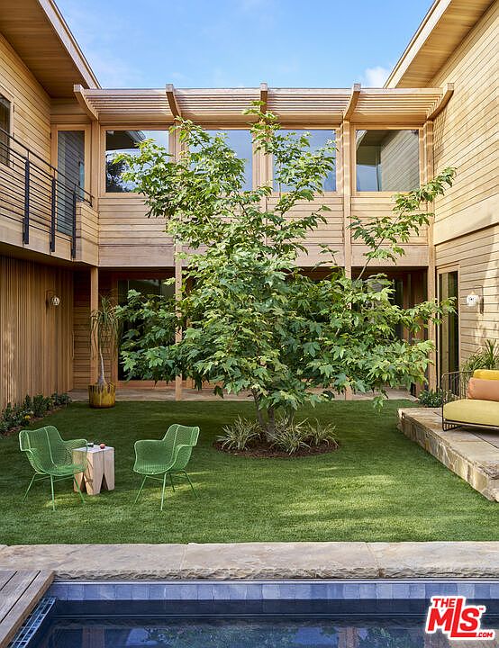 This serene courtyard features a lush green lawn centered around a mature tree, framed by modern light-wood siding and large windows. Two vibrant green wire chairs sit beside a minimalist wooden side table, creating a peaceful outdoor seating area. The foreground includes the edge of a swimming pool, emphasizing a seamless transition between indoor and outdoor living spaces.