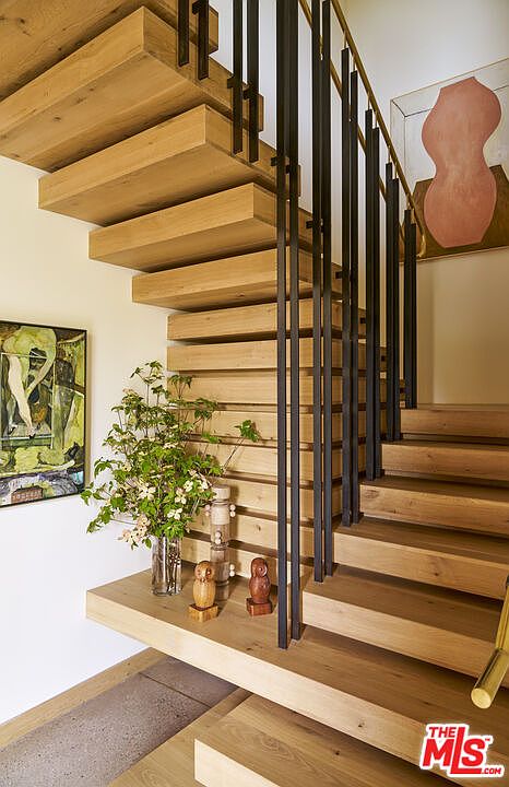 This image captures a striking staircase featuring floating light wood treads and a minimalist black metal railing. The design is complemented by abstract artwork on the walls and a decorative arrangement of greenery and small sculptures on the landing. The perspective is from the base of the stairs, looking up to emphasize the clean lines and modern architectural aesthetic.