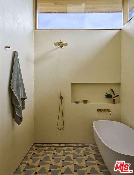 This minimalist bathroom features a wet-room style layout with a freestanding white soaking tub and a wall-mounted shower head. The space is finished in smooth, neutral-toned plaster walls and accented by a geometric patterned tile floor, while a clerestory window above provides natural light. A recessed wall niche offers built-in storage, and brass-toned fixtures add a touch of warmth to the clean, modern aesthetic.
