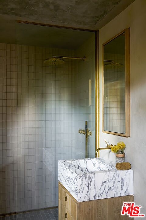 This modern bathroom features a striking stone vessel sink atop a light wood vanity, complemented by a gold-toned faucet. To the left, a glass-enclosed shower area showcases a grid-patterned tile wall and matching gold fixtures. The space is finished with a textured ceiling and a framed mirror, creating a sophisticated and contemporary aesthetic.
