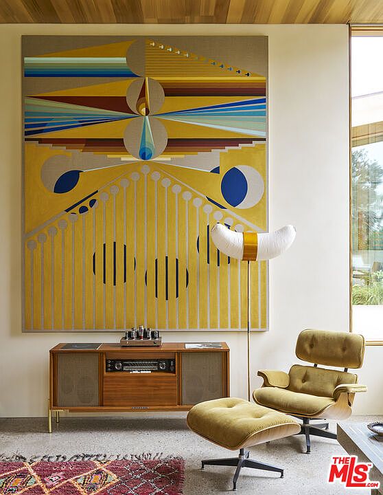 This interior space features a mid-century modern aesthetic, highlighted by a large, vibrant geometric art piece mounted on the wall above a vintage-style wooden media console. To the right, a classic lounge chair with an ottoman sits beside a unique, crescent-shaped floor lamp, creating a cozy reading nook. The room is bathed in natural light from a tall window, and the scene is anchored by a textured, patterned rug in the foreground.