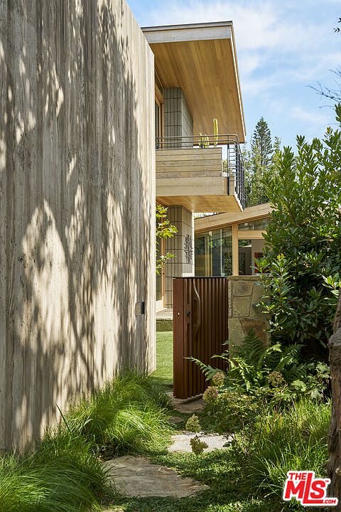 This side view captures a modern architectural design featuring a prominent, textured concrete wall that creates a dramatic play of light and shadow. A wooden balcony extends from the upper level, complemented by a vertical, slatted metal gate that leads toward the rear of the property. Lush greenery, including ornamental grasses and shrubs, frames the stone-paved path, enhancing the home's contemporary and organic aesthetic.