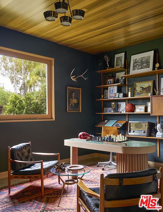 This sophisticated home office features dark-painted walls contrasted by a warm, light wood-paneled ceiling and a matching wood-framed window. The room is furnished with a unique, curved-edge desk, two mid-century modern leather armchairs, and a glass-topped coffee table set atop a patterned area rug. A large, multi-tiered shelving unit displays various collectibles, books, and art, creating a curated and scholarly atmosphere.
