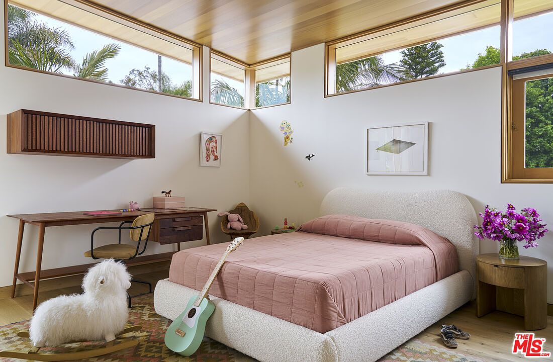 This bright and airy bedroom features a textured, light-colored bed frame paired with soft pink bedding, creating a cozy and inviting atmosphere. The room is accented by a wooden desk with a matching chair, a wall-mounted storage unit, and a light-colored wood ceiling that adds warmth to the space. Large windows wrap around the corner of the room, providing ample natural light and views of the surrounding greenery.