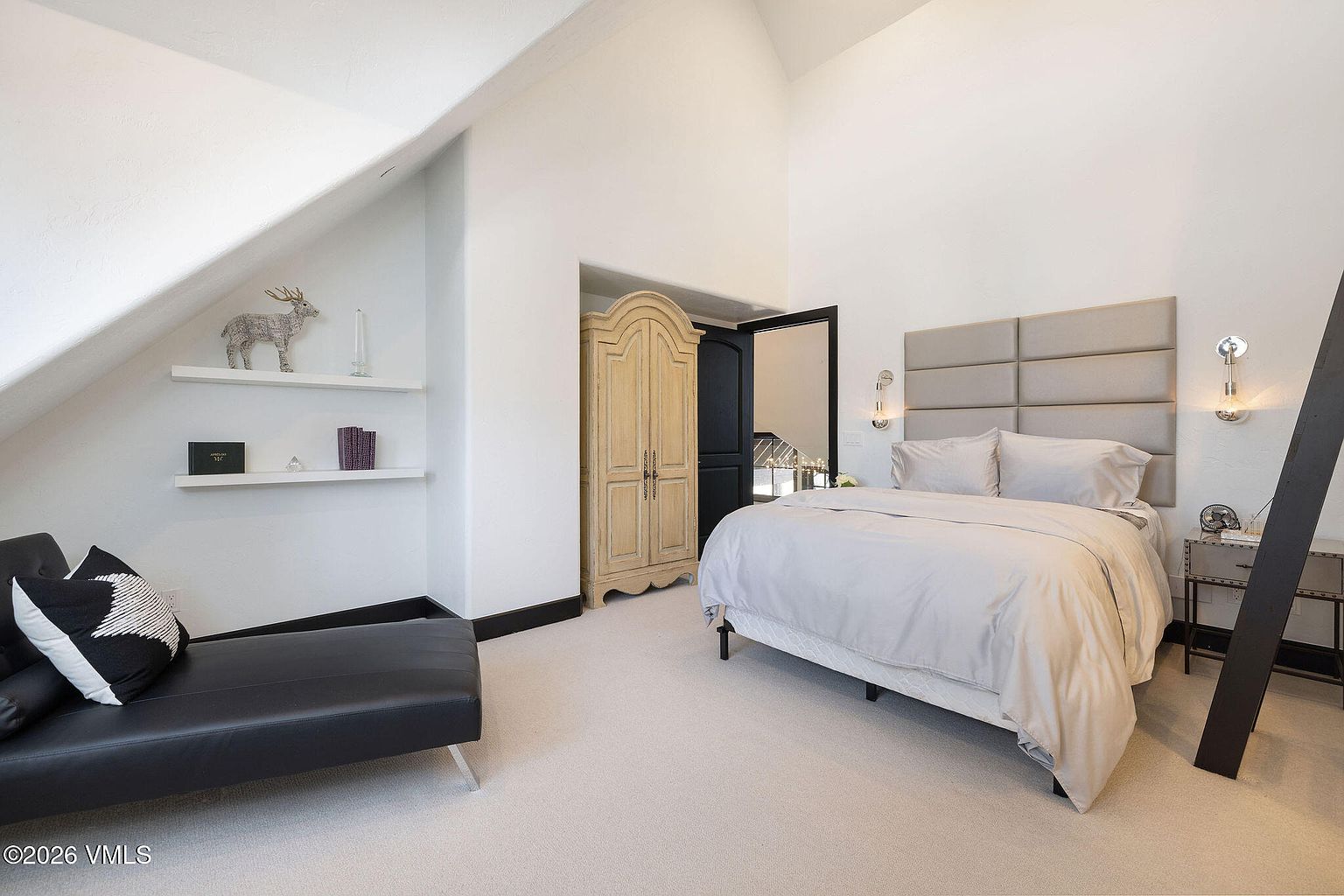 This is a bright and airy primary bedroom featuring a neutral color palette. The room includes a large bed with a padded headboard, a black leather chaise lounge, and a light wood armoire. The high ceilings and minimalist decor create a serene and spacious atmosphere, perfect for relaxation.