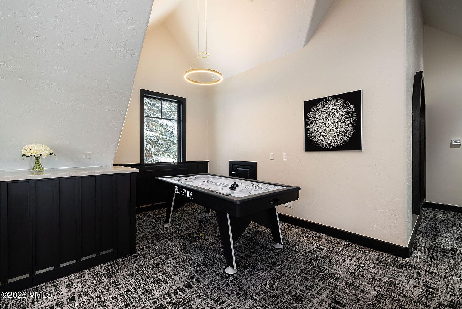This interior shot showcases a game room featuring an air hockey table as the focal point, illuminated by a modern ring chandelier. The room is decorated with a black and white color scheme, including a striking piece of wall art and a patterned carpet. A window provides natural light, and the overall impression is a stylish and inviting space for entertainment.