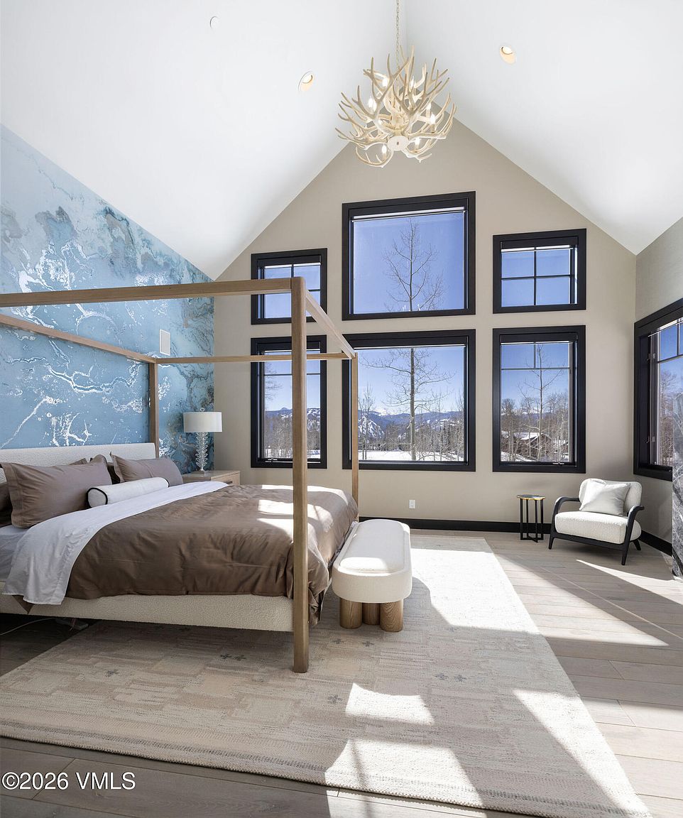 This is a bright and airy primary bedroom featuring a four-poster bed with neutral bedding, a statement blue marble-esque accent wall, and a unique antler chandelier. Large windows offer stunning mountain views, enhancing the room's serene and luxurious atmosphere. The room is decorated in a modern style with a neutral color palette and natural wood accents.