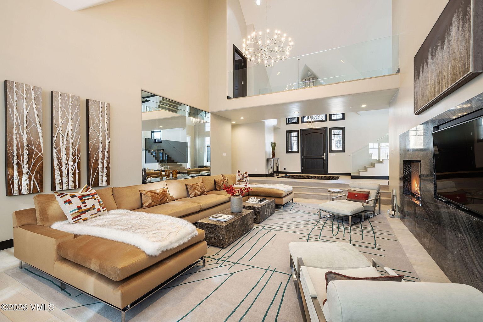 This is a spacious living room featuring a large sectional sofa with decorative pillows and a faux fur throw. The room has high ceilings, a modern chandelier, and artwork depicting birch trees. A fireplace is visible on the right, and the overall style is contemporary and luxurious.