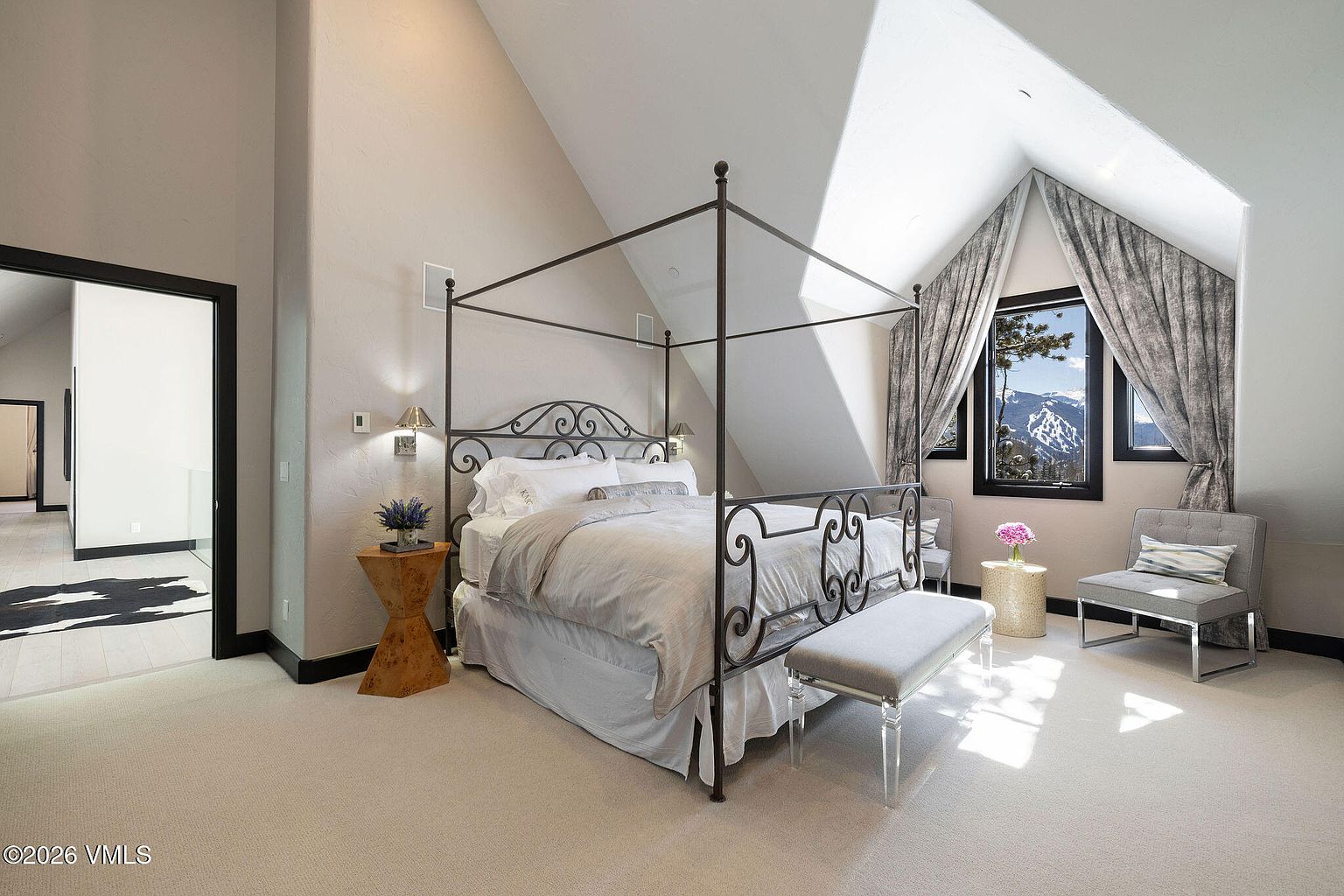 This is a primary bedroom featuring a four-poster bed with wrought iron detailing, creating a luxurious and elegant atmosphere. The room has an angled ceiling with a window that offers a view of the mountains. The color palette is neutral, with soft lighting and plush carpeting, contributing to a serene and inviting space.