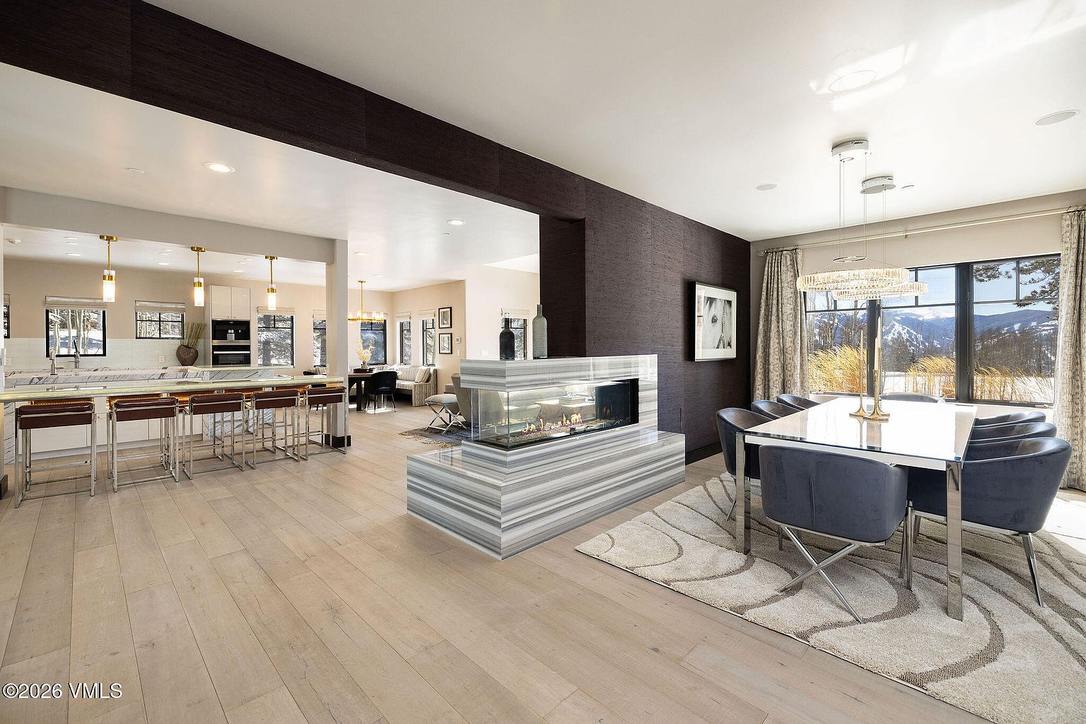 This interior shot showcases a modern dining area adjacent to a kitchen and living space. The dining room features a glass-topped table with chrome legs, surrounded by plush gray chairs, and illuminated by a contemporary chandelier. A unique, see-through fireplace separates the dining area from the living room, adding a touch of modern elegance.