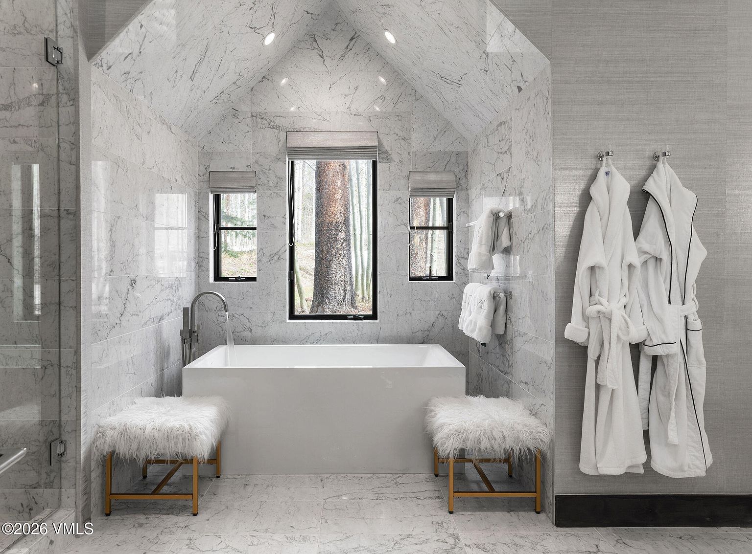 This is a luxurious primary bathroom featuring marble walls and flooring, a freestanding white bathtub positioned beneath a large window offering a view of trees, and two plush, furry stools on either side of the tub. Two white robes hang on the right wall, and the overall impression is one of spa-like tranquility and high-end design.