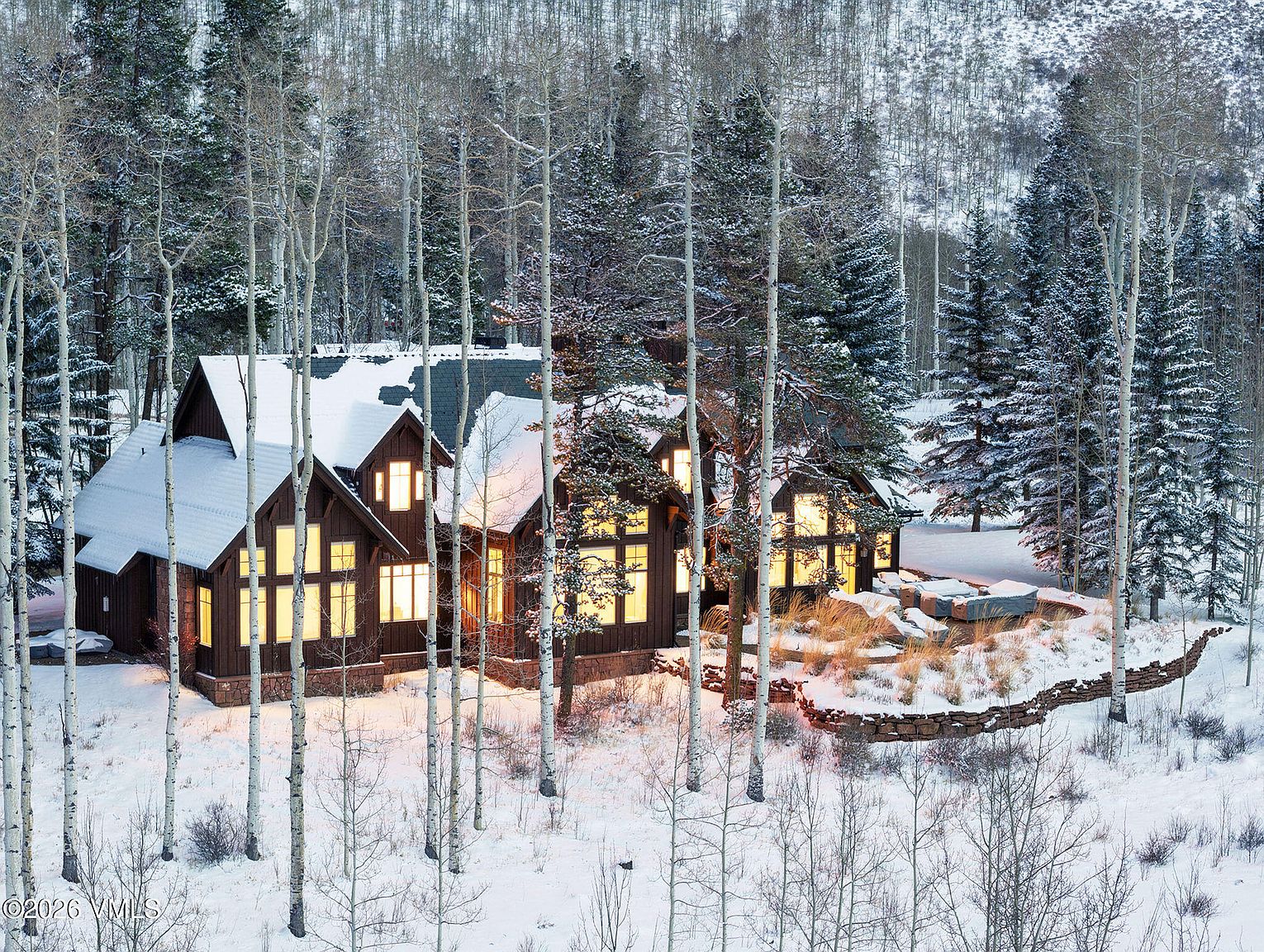 This image showcases the front exterior of a beautiful mountain home in a snowy, wooded setting. The home features a dark wood facade, multiple windows with warm interior lighting, and a roof partially covered in snow. Tall, bare trees in the foreground add to the wintery ambiance, creating a cozy and inviting scene.