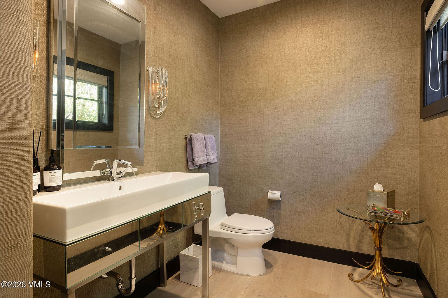 This is a well-appointed guest bathroom featuring textured wallpaper, a modern rectangular sink with a mirrored vanity, and a toilet. The room is decorated with a glass side table and neutral-toned accessories, creating a sophisticated and clean aesthetic. The flooring appears to be light wood, complementing the overall color scheme.