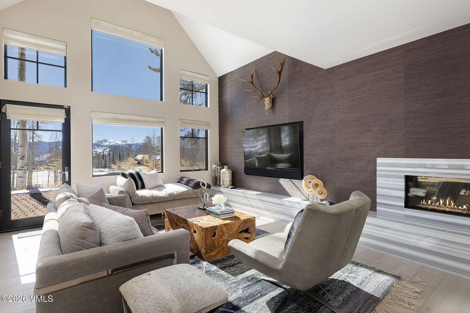 This is an interior shot of a modern living room featuring a high ceiling and large windows that offer a view of a snowy landscape. The room is furnished with a gray sofa, a unique wooden coffee table, and a stylish armchair, complemented by a textured rug. A fireplace and a mounted television add to the room's appeal, creating a cozy and sophisticated atmosphere.