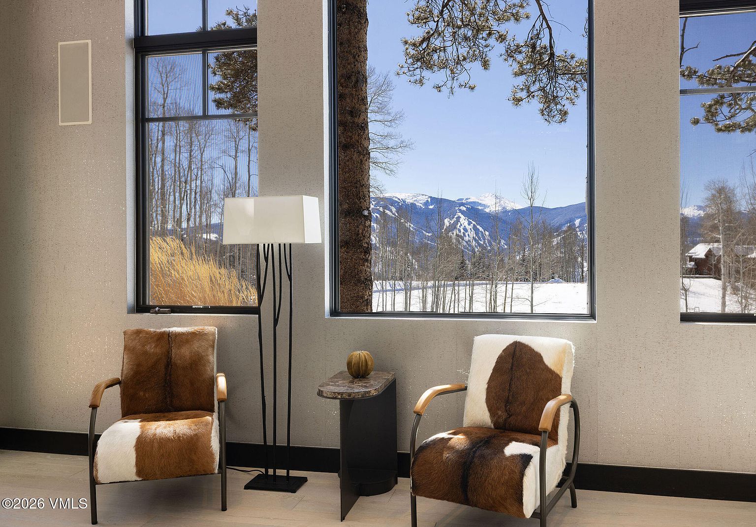 This interior shot showcases a cozy living room area with two unique cowhide chairs flanking a modern floor lamp and a small side table. Large windows offer a stunning view of a snow-covered landscape with mountains in the distance, creating a serene and inviting atmosphere. The room's design blends rustic elements with contemporary style, perfect for a mountain retreat.