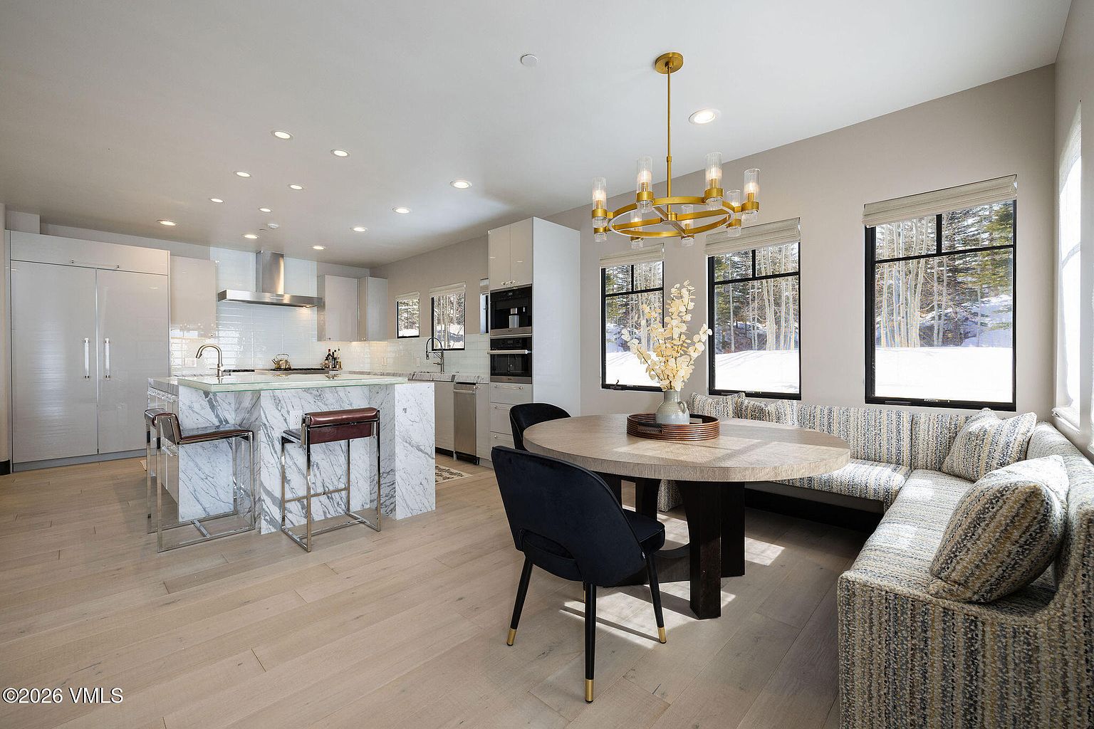This interior shot showcases a modern kitchen and dining area. The kitchen features sleek white cabinetry, stainless steel appliances, and a marble-clad island with bar seating. The dining area includes a round table with black chairs and a built-in banquette seating area, illuminated by a stylish gold chandelier, creating a bright and inviting space.