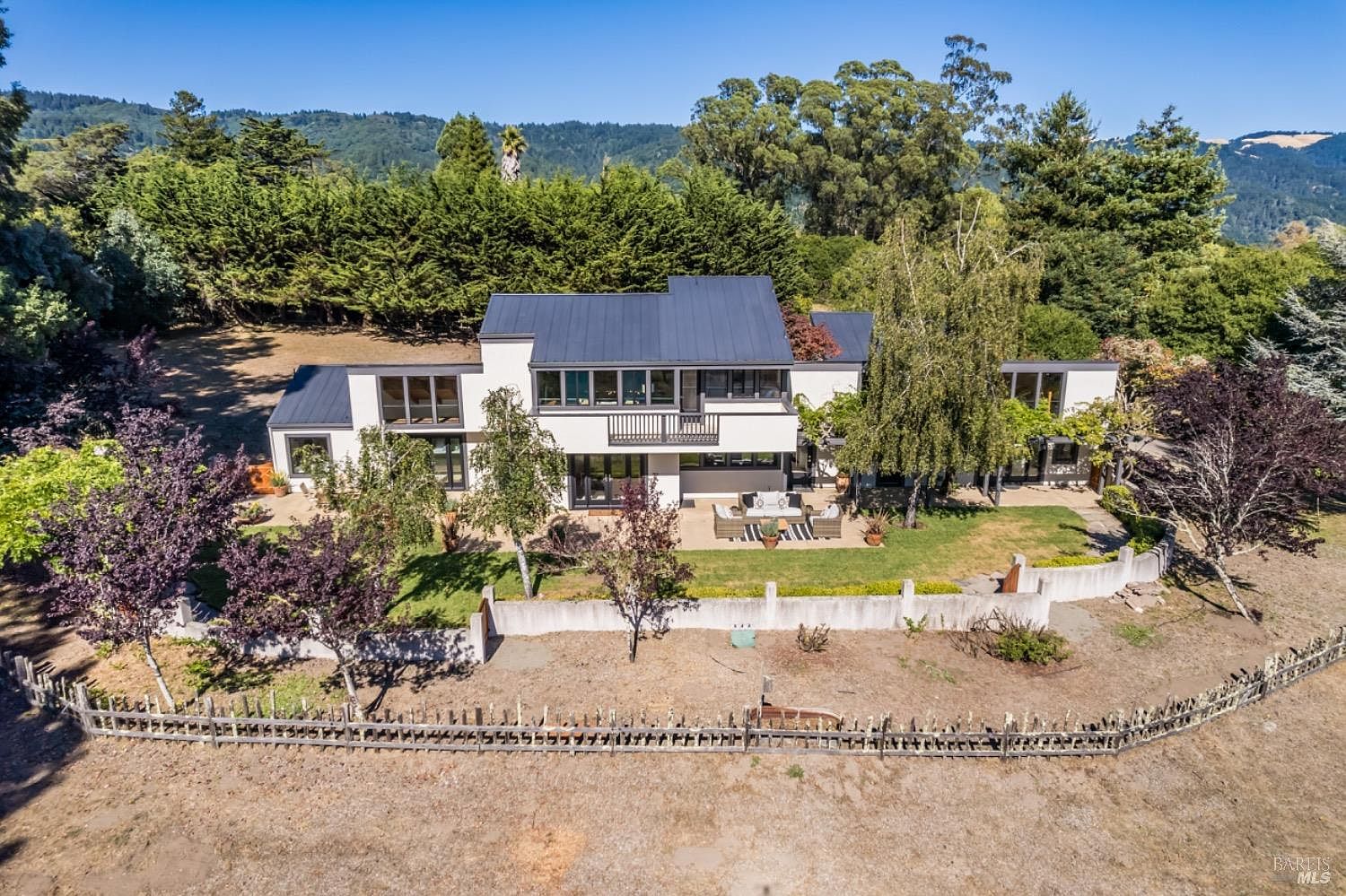 This aerial perspective captures a modern, multi-level residence nestled against a backdrop of lush, rolling hills and dense forest. The home features a striking dark metal roof, clean white stucco walls, and a spacious patio area furnished for outdoor living, all enclosed by a low concrete wall and a rustic wooden fence. The composition highlights the property's serene, private setting and its seamless integration with the surrounding natural landscape.