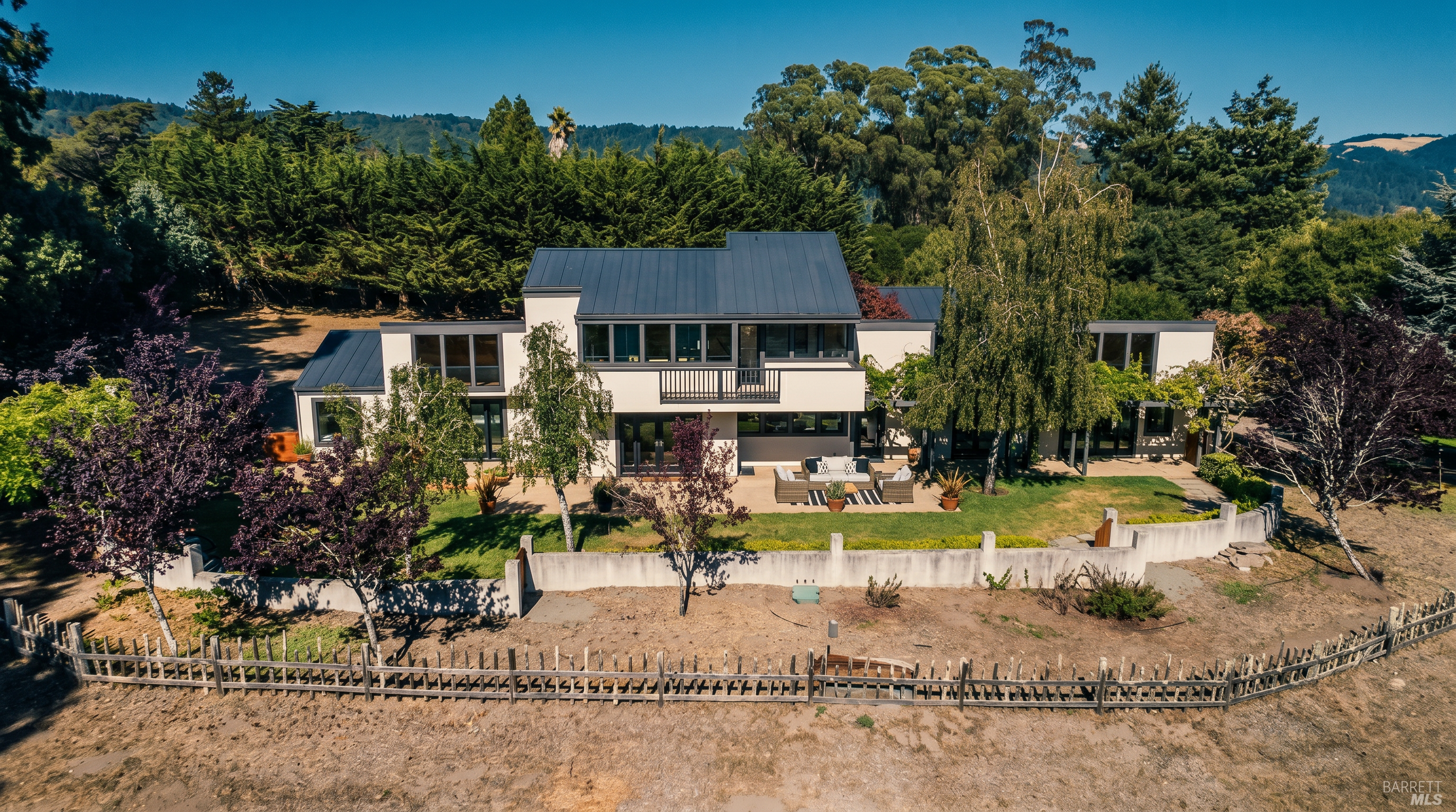This aerial perspective captures a modern, multi-level residence nestled against a backdrop of lush, rolling hills and dense forest. The home features a striking dark metal roof, clean white stucco walls, and a spacious patio area furnished for outdoor living, all enclosed by a low concrete wall and a rustic wooden fence. The composition highlights the property's serene, private setting and its seamless integration with the surrounding natural landscape.