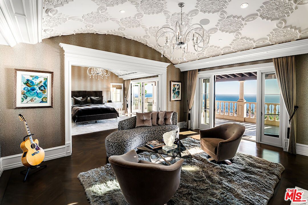 This is an elegant living room featuring a unique ceiling design with intricate patterns and a chandelier. The room opens to a bedroom on one side and a balcony with ocean views on the other, creating a luxurious and airy atmosphere. The decor includes a stylish chaise lounge, plush rug, and tasteful artwork, enhancing the room's sophisticated appeal.