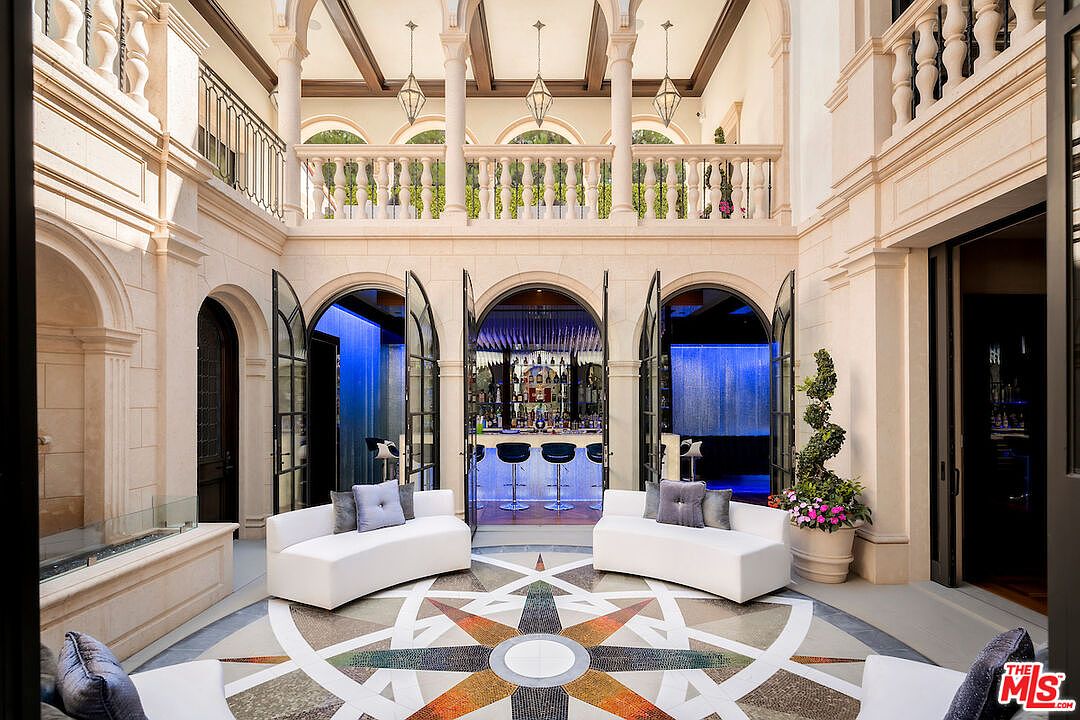 This grand interior space features a stunning mosaic floor design, complemented by two curved white sofas with gray pillows. Arched doorways lead to a bar area and a waterfall feature, enhancing the luxurious ambiance. The space is bathed in natural light, highlighting the architectural details and creating an inviting atmosphere.