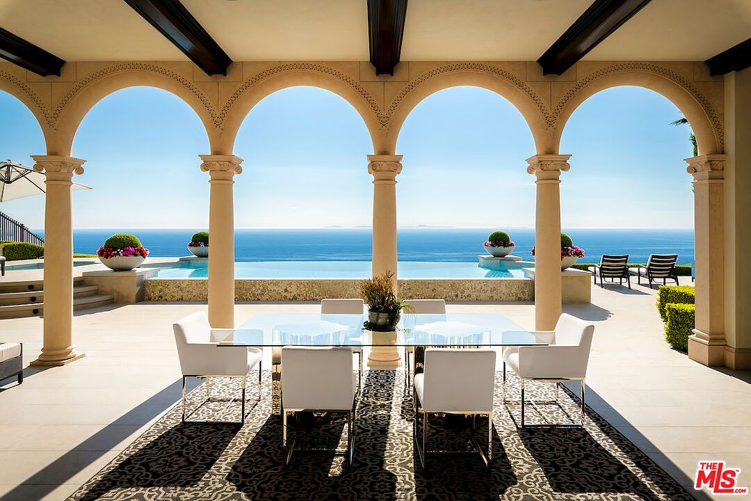 This image showcases a luxurious outdoor patio area with a stunning ocean view. The patio features a dining table with white chairs, set on a patterned rug, and is framed by elegant arches and columns. Beyond the patio, there's a pool and lounge chairs, creating an inviting space for relaxation and entertainment.
