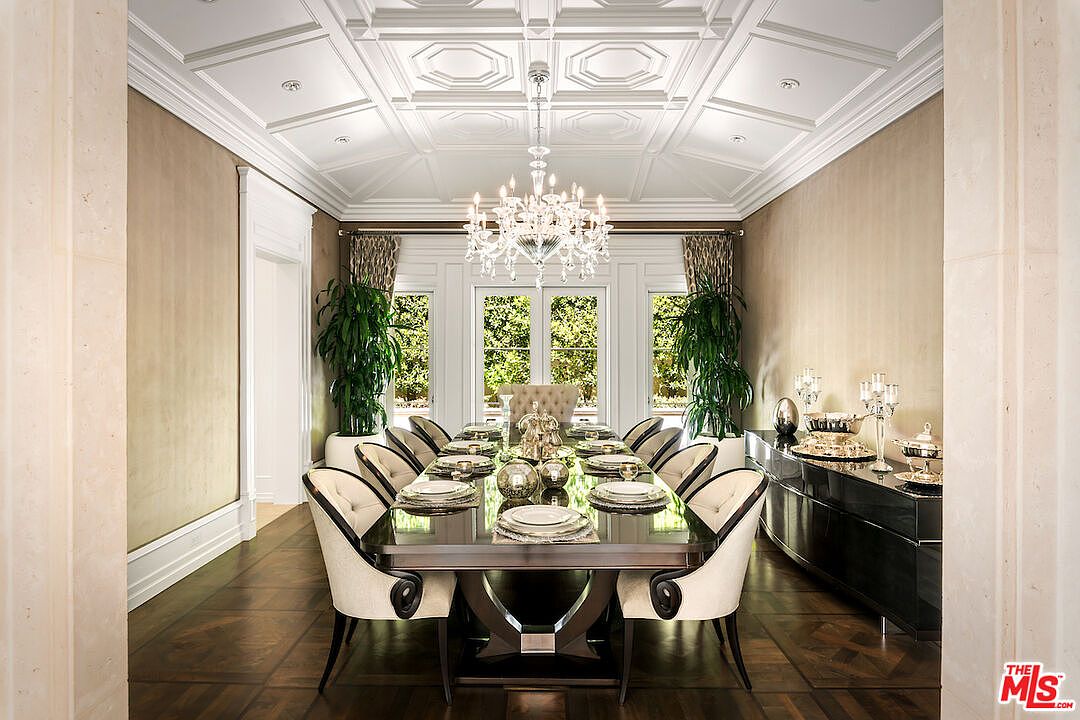 This is an elegant dining room featuring a long, dark wood table set for a formal meal, surrounded by upholstered chairs with dark wood frames. A crystal chandelier hangs above the table, illuminating the room, and large windows with patterned curtains provide natural light and a view of the outdoors. A dark wood sideboard is placed against the wall, adding to the room's sophisticated ambiance.