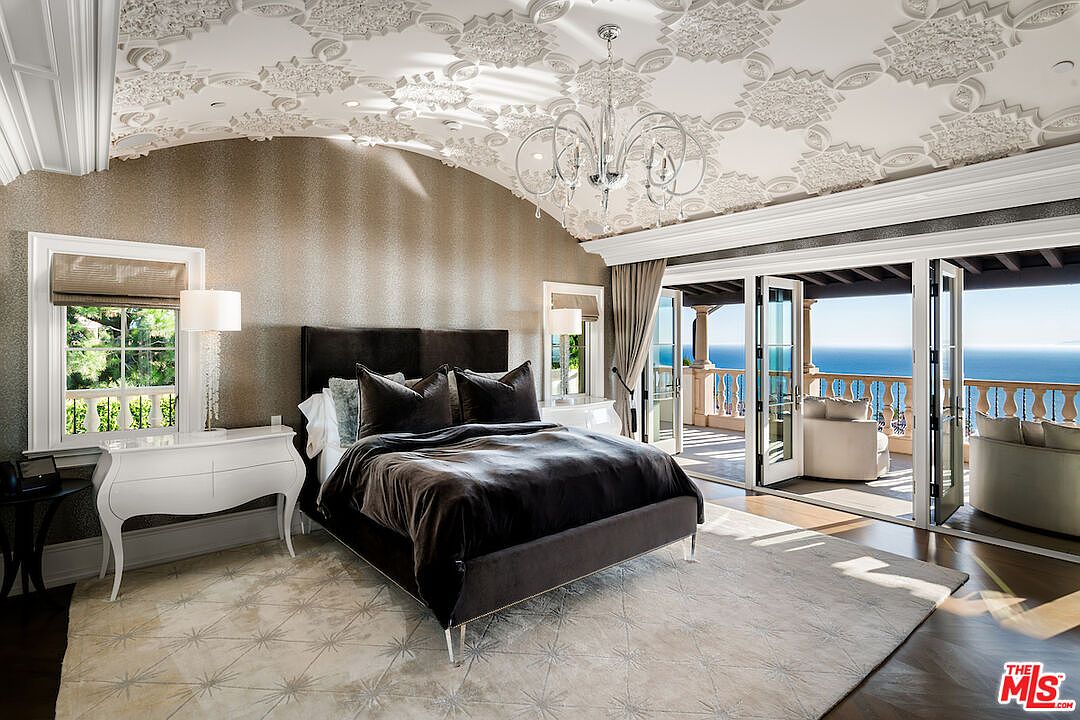 This is a luxurious primary bedroom featuring a dark upholstered bed with velvet bedding, complemented by a light-colored rug and a white dresser. The room boasts an arched ceiling with intricate detailing and a chandelier, while large glass doors open onto a balcony with ocean views. The perspective is from the foot of the bed, showcasing the room's depth and connection to the outdoor scenery.
