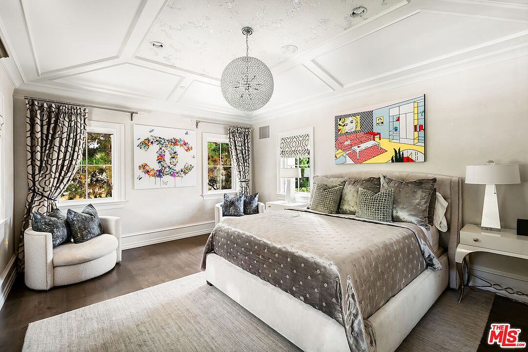 This is a luxurious primary bedroom featuring a large bed with a tufted headboard and plush bedding. The room is decorated with modern art, a crystal chandelier, and custom window treatments. The overall style is elegant and sophisticated, creating a relaxing and inviting atmosphere.