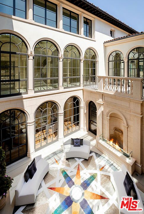 This image showcases a luxurious outdoor patio area with a unique starburst-patterned tile floor. The space is furnished with modern white lounge seating and features a built-in fireplace, creating a sophisticated and inviting atmosphere. Large arched windows provide ample natural light and views of the surrounding architecture.