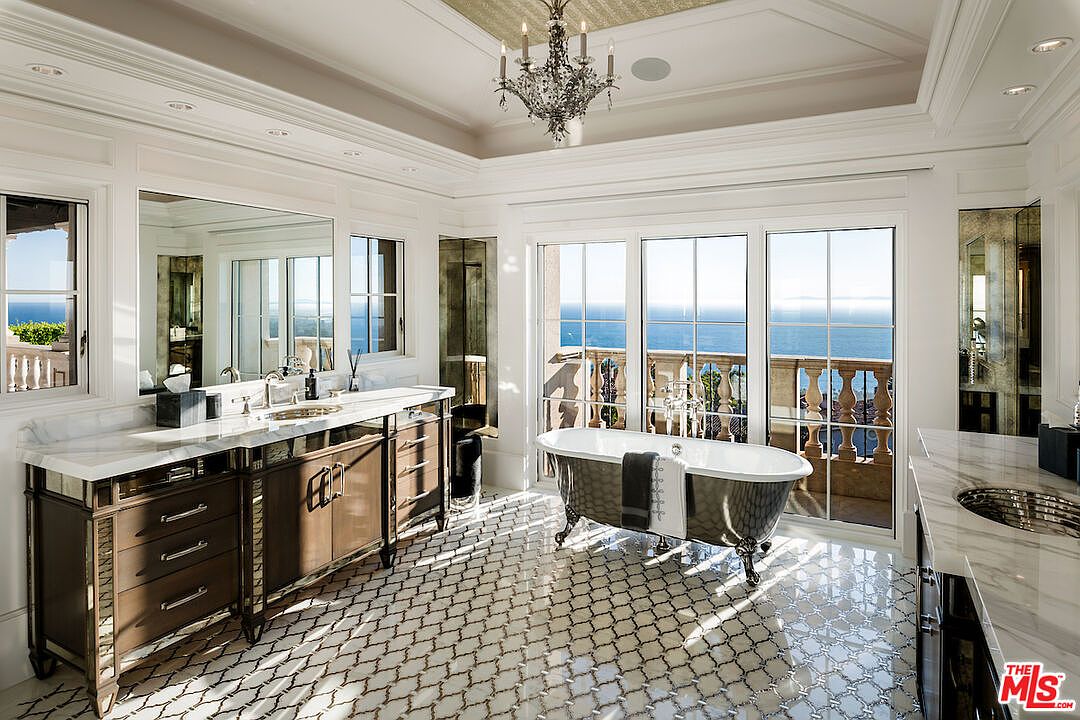 This is a luxurious primary bathroom featuring a freestanding bathtub positioned in front of large windows with ocean views. The room is adorned with elegant fixtures, including a chandelier, and a vanity with a marble countertop. The patterned tile flooring adds a touch of sophistication to the space.