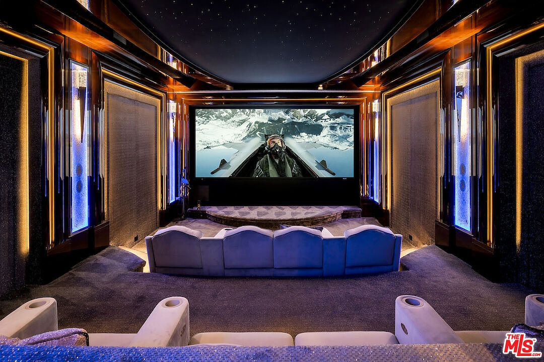This is a luxurious home theater featuring tiered seating with plush, velvet-like chairs facing a large screen displaying a scene from a movie. The room is designed with dark, rich wood paneling accented by custom lighting, creating an immersive cinematic experience. The ceiling is adorned with star-like lights, enhancing the theater's ambiance.