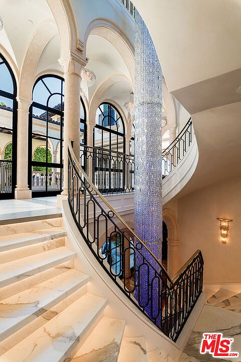 This interior shot showcases a grand, curved staircase with marble steps and an ornate black iron railing accented with gold. A massive, cylindrical chandelier hangs in the center, adding a dramatic focal point. Arched windows and doorways provide natural light and architectural interest, creating a luxurious and elegant ambiance.