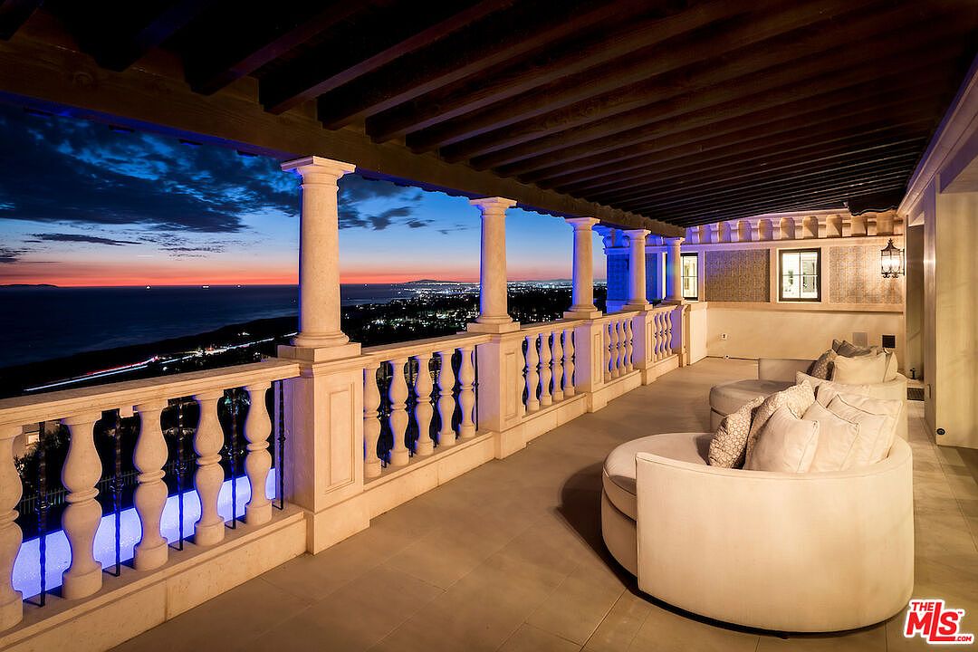 This image showcases a luxurious outdoor balcony or patio area with elegant architectural details, including stone columns and balustrades. Comfortable, rounded seating arrangements invite relaxation, while the stunning sunset and city lights provide a breathtaking backdrop. The space exudes sophistication and offers an ideal setting for outdoor entertaining or quiet contemplation.