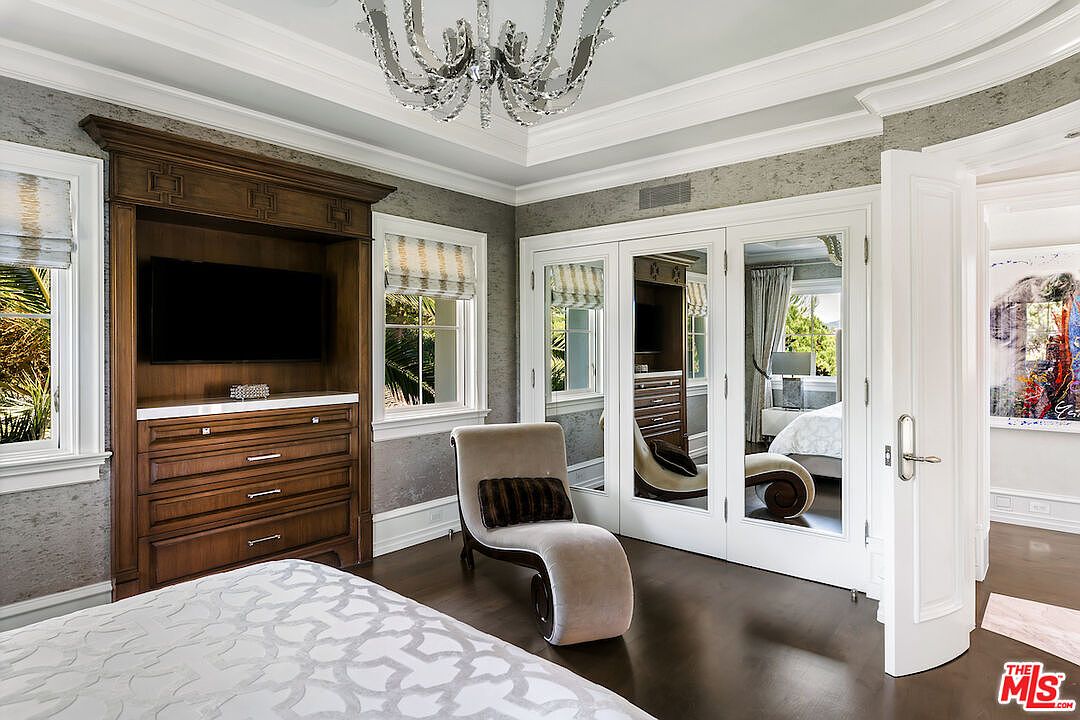This primary bedroom features a built-in entertainment center with a dark wood finish, a flat-screen TV, and drawers for storage. A chaise lounge sits near mirrored closet doors, reflecting the room's elegant decor. The room is illuminated by a crystal chandelier, and the walls are adorned with textured wallpaper, creating a luxurious and comfortable atmosphere.