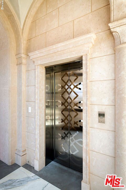 The image showcases an interior architectural detail, specifically an elevator framed by ornate stone archways and columns. The elevator doors feature a geometric design, adding a modern touch to the classic architectural style. The overall impression is one of luxury and sophisticated design.