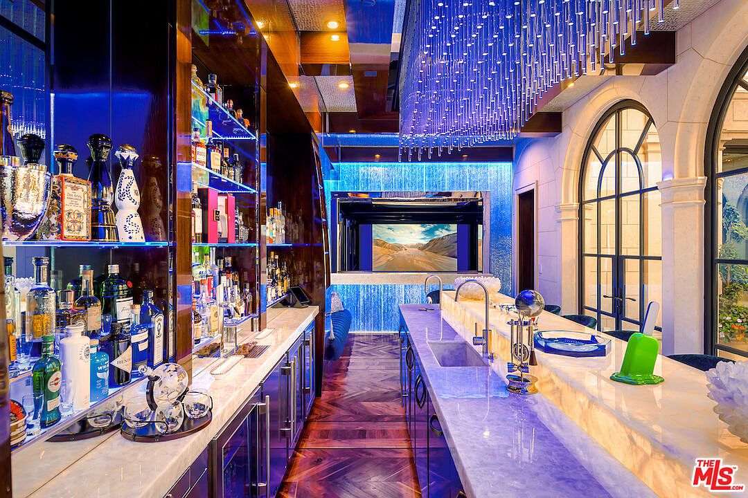 This is a luxurious wine cellar featuring a well-stocked bar with illuminated shelving displaying various bottles. The bar area includes a marble countertop with sinks and a unique chandelier overhead, complemented by a waterfall feature behind a large screen displaying a desert landscape. The flooring is a rich, dark wood in a herringbone pattern, adding to the sophisticated ambiance.
