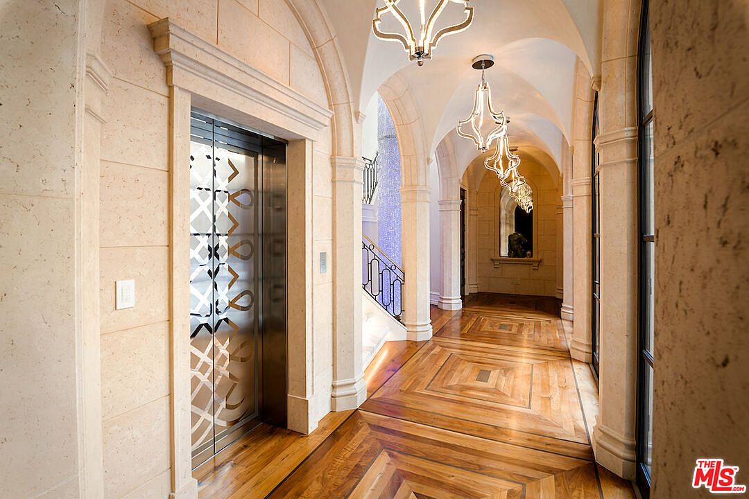 This interior shot showcases a grand hallway with arched ceilings and elegant chandeliers. The hallway features a patterned hardwood floor and stone walls, leading to a staircase with ornate railings. An elevator with decorative doors is visible on the left, adding a touch of modern convenience to the classic design.